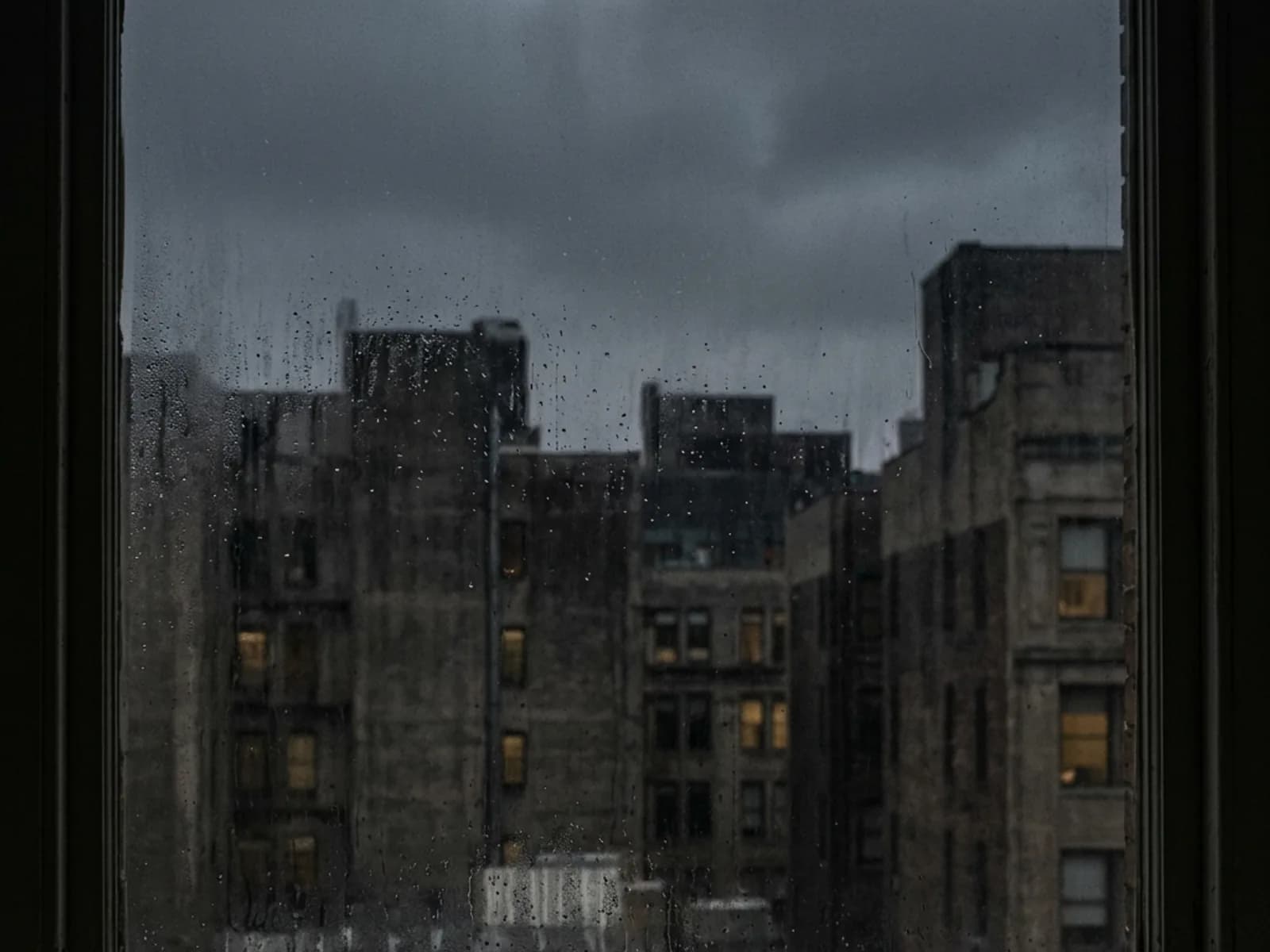 City buildings at dusk seen through a rain-speckled window, a few windows lit.